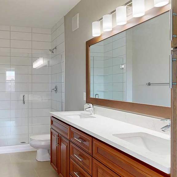 Thumbnail 23 of 27 A bathroom with a cherry-stained wooden vanity with white quartz countertops, and a tiled walk-in shower with a glass door.