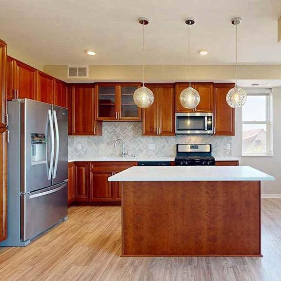 Thumbnail 10 of 27 A kitchen with cherry-stained wooden cabinets, light quartz countertops, stainless steel appliances, and an island with pendant lighting.