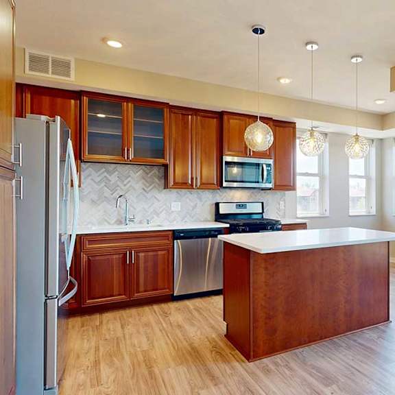 Thumbnail 13 of 27 A kitchen with cherry-stained wooden cabinets, light quartz countertops, stainless steel appliances, and an island with pendant lighting.