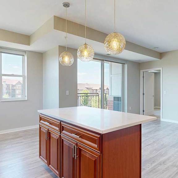 Thumbnail 17 of 27 A kitchen island with cherry-stained wooden cabinets and a light quartz countertop, in front of a spacious living room.