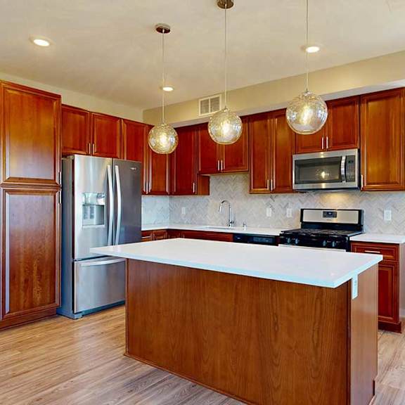 Thumbnail 11 of 29 A kitchen with cherry-stained wooden cabinets, light quartz countertops, stainless steel appliances, and an island with pendant lighting.