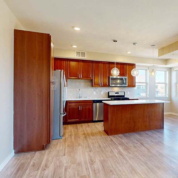 Thumbnail 7 of 29 A kitchen with cherry-stained wooden cabinets, light quartz countertops, stainless steel appliances, and an island with pendant lighting in front of a spacious living room.
