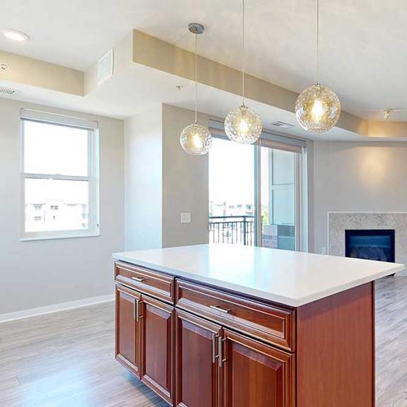 Thumbnail 16 of 29 A kitchen island with cherry-stained wooden cabinets and a light quartz countertop, in front of a spacious living room with a fireplace.
