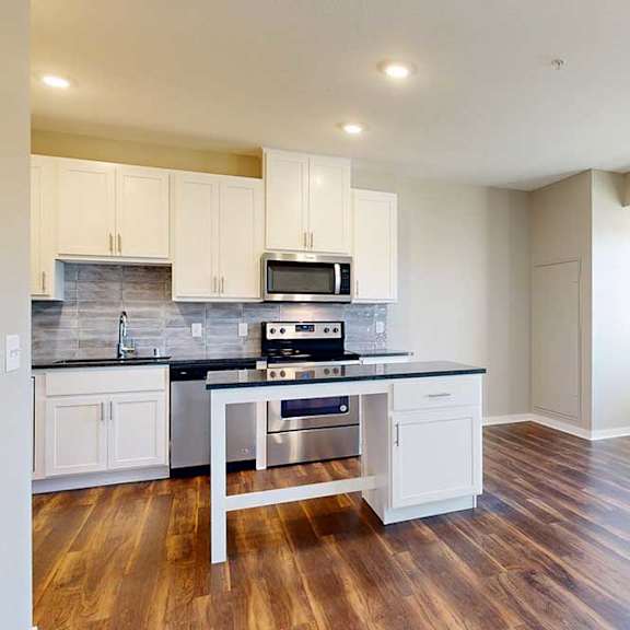 Thumbnail 7 of 13 A modern kitchen with white cabinets and an island, open to a living area with large windows and hardwood-style flooring.