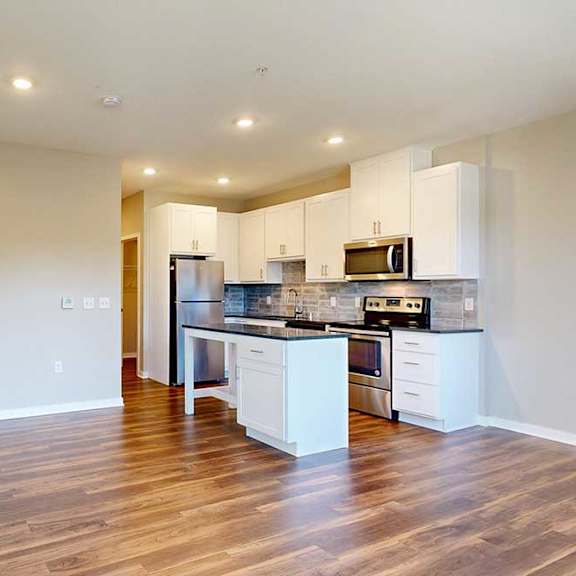 Thumbnail 10 of 13 A living area with hardwood-style flooring, open to a modern kitchen with white cabinets and an island.