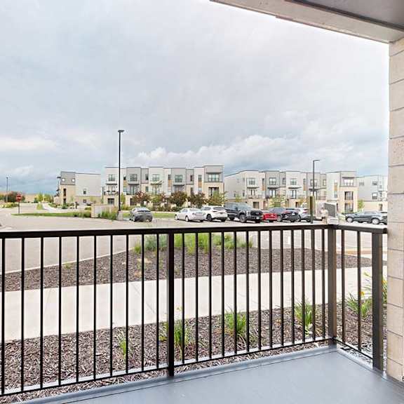 Thumbnail 5 of 19 A balcony with a black railing overlooks a parking lot and apartment buildings.