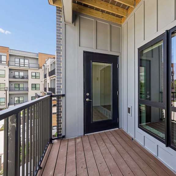 Thumbnail 7 of 17 Private apartment balcony with wood-look decking, black railing, and views of the community buildings and surrounding neighborhood.