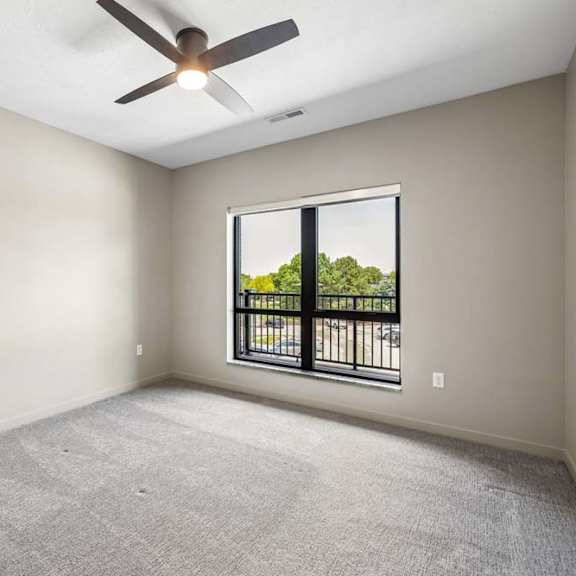 Thumbnail 13 of 17 Carpeted apartment bedroom with a ceiling fan, large window for natural light, and neutral wall finishes throughout.