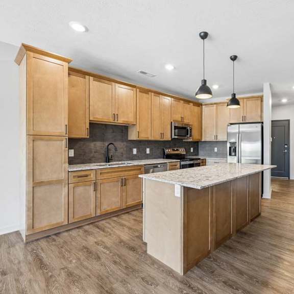 Thumbnail 4 of 17 Open-concept apartment kitchen with wood cabinetry, a center island, pendant lighting, and wood-look flooring extending into the living area.