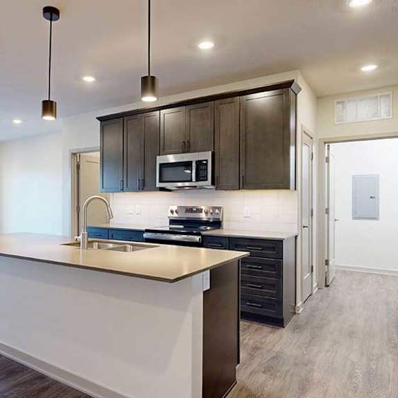 Thumbnail 9 of 24 A kitchen with dark wooden cabinets and a large island, in front of a spacious living room with floor-to-ceiling windows.