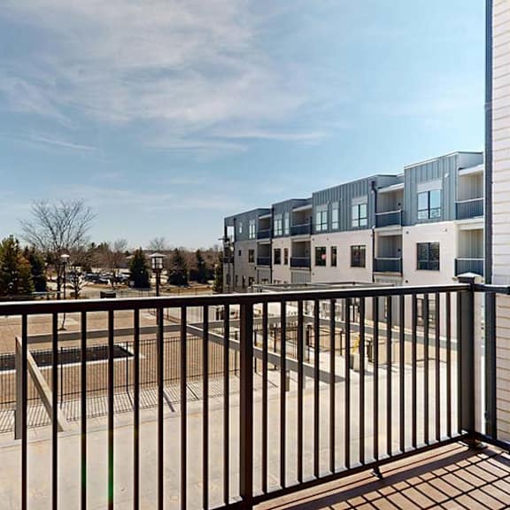 Thumbnail 42 of 42 Private apartment balcony with black metal railing overlooking the community courtyard and neighboring residential buildings under a clear blue sky.