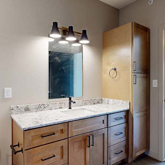 Thumbnail 40 of 42 Modern apartment bathroom with a single-sink vanity, wood cabinetry, a stone-look countertop, large mirror, vanity lighting, and an adjoining bedroom visible through the open doorway.