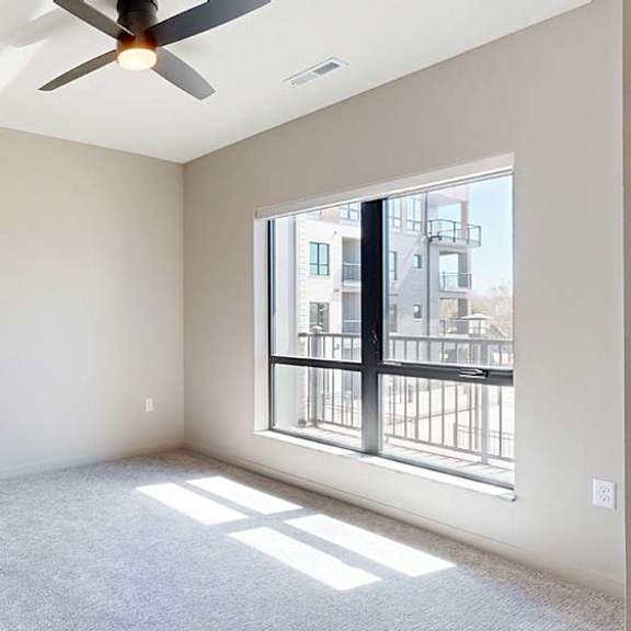 Thumbnail 38 of 42 Bright apartment bedroom with soft carpeting, a large window providing natural light, a ceiling fan, neutral finishes, and views of the neighboring community buildings.