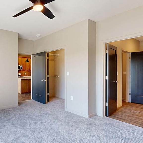 Thumbnail 36 of 42 Bright apartment bedroom with soft carpeting, a large window, ceiling fan, neutral walls, and an open doorway leading toward the kitchen and hallway.