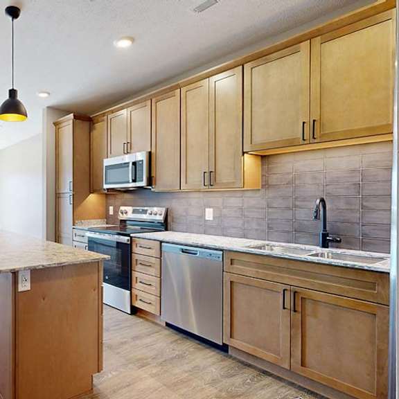 Thumbnail 31 of 42 Bright apartment kitchen with wood cabinetry, a large island, tile backsplash, stainless steel appliances, pendant lighting, and an open layout connecting to the living area.