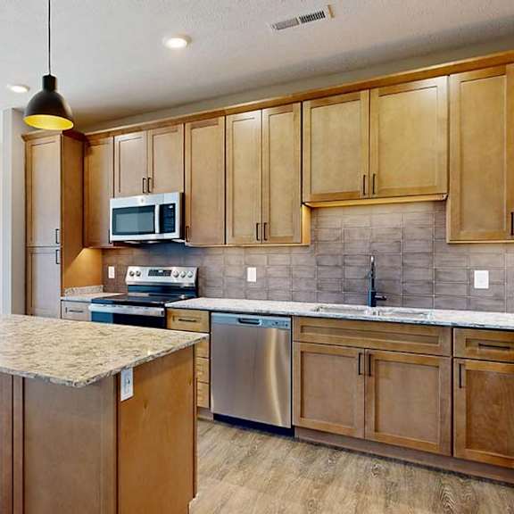 Thumbnail 32 of 42 Spacious apartment kitchen with wood cabinetry, a large island, tile backsplash, stainless steel appliances, pendant lighting, and an open layout with adjacent living space and large windows.