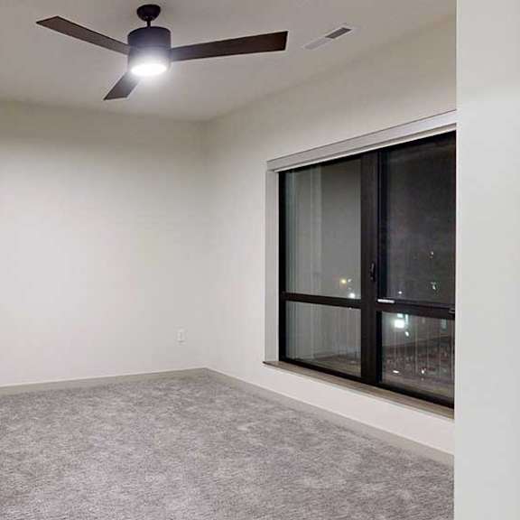 Thumbnail 75 of 84 Bright apartment bedroom with neutral carpeting, a ceiling fan, and a large window overlooking nighttime views.