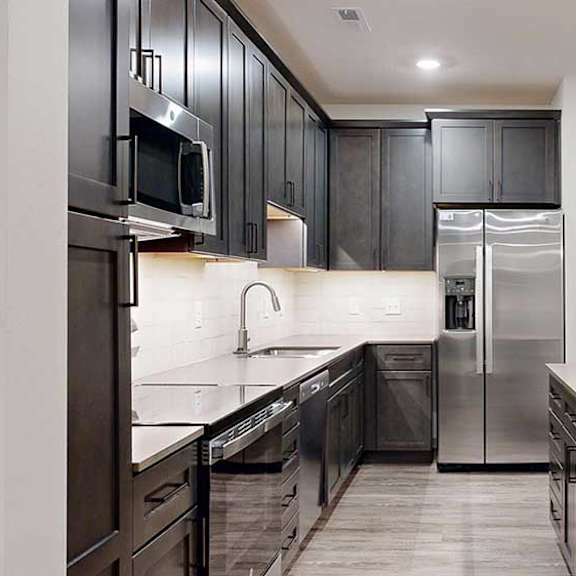 Thumbnail 72 of 84 Modern apartment kitchen with dark cabinetry, stainless steel appliances, light countertops, and a large island with pendant lighting.
