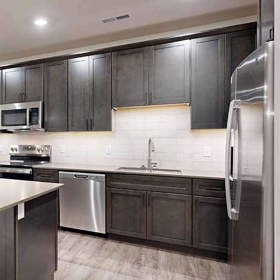 Thumbnail 71 of 84 Modern apartment kitchen with dark cabinetry, stainless steel appliances, a large island, and pendant lighting.