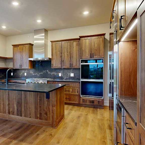 Thumbnail 17 of 46 A gourmet kitchen with an oversized island, oak wooden cabinetry, stainless steel commercial appliances, dark quartz countertops, a range hood, and a tile backsplash.