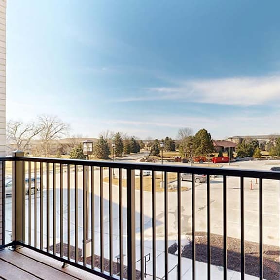 Thumbnail 46 of 46 Private apartment balcony with black metal railing overlooking a courtyard, neighboring buildings, trees, and open blue sky.