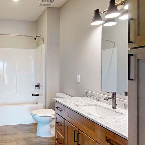 Thumbnail 43 of 46 Modern apartment bathroom with a quartz-style vanity, wood cabinetry, a large mirror, and a tub-shower combination with a sliding glass shower door.