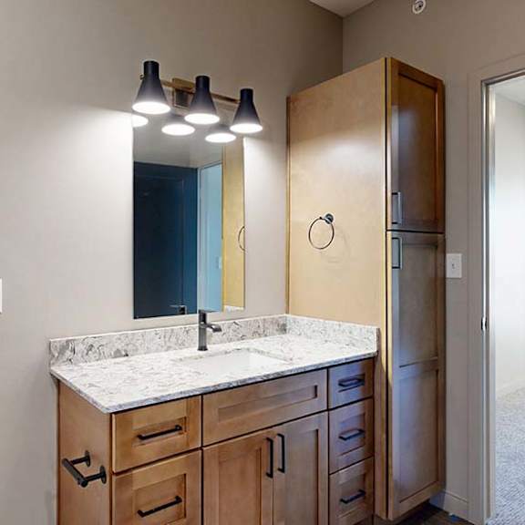 Thumbnail 45 of 46 Modern apartment bathroom with a quartz-style vanity, wood cabinetry, contemporary lighting, and a built-in linen cabinet, with the adjacent bedroom visible through the doorway.