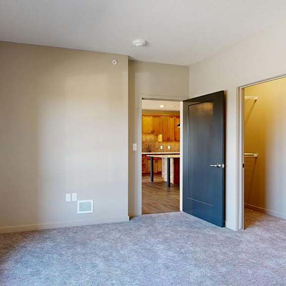 Thumbnail 38 of 46 Bright apartment bedroom with neutral carpeting, a large window for natural light, and an open doorway leading to the adjacent living area and kitchen.