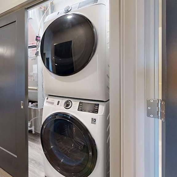 Thumbnail 37 of 46 Apartment laundry closet with a stacked front-load washer and dryer, conveniently located off the kitchen with wood-look flooring and modern interior finishes.