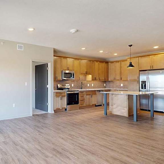 Thumbnail 36 of 46 Bright open-concept apartment living room with wood-look flooring, large windows, and a modern kitchen featuring a center island, wood cabinetry, and pendant lighting in the background.