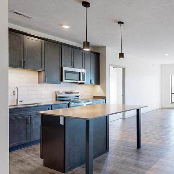 Thumbnail 8 of 30 a modern kitchen with a table island and dark cabinets next to a living room with two large windows