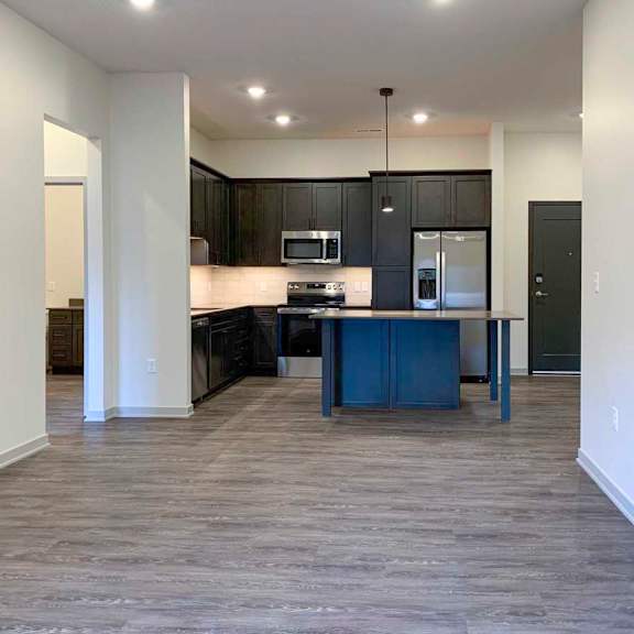Thumbnail 15 of 29 a living room with wooden flooring in front of a kitchen with dark cabinets and a large center island