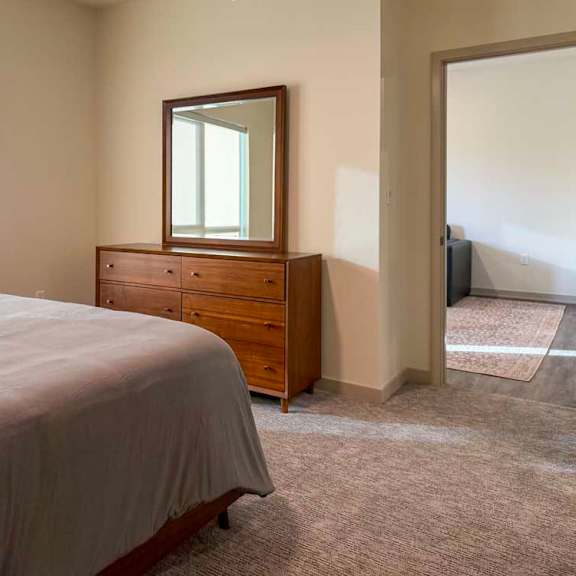Thumbnail 27 of 42 Bright bedroom with carpet flooring, large window, bed, and wood dresser with mirror, opening to an adjacent living area.