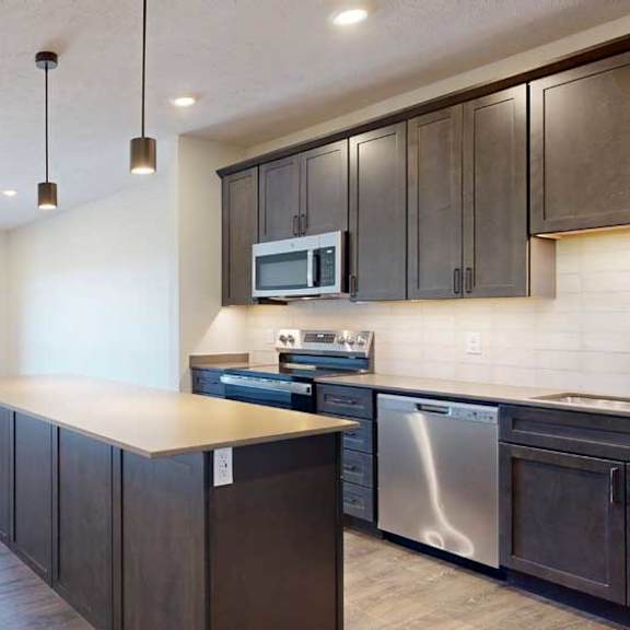 Thumbnail 2 of 21 A kitchen with dark wooden cabinets and a large island, in front of a spacious living room with floor-to-ceiling windows.