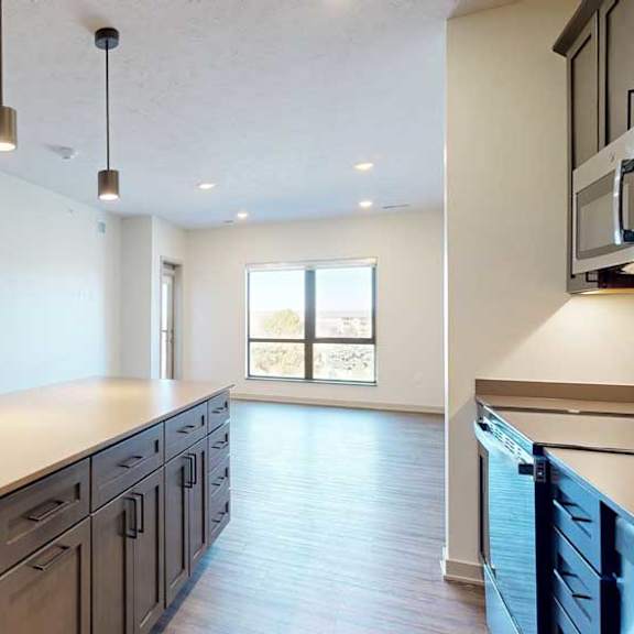 Thumbnail 7 of 21 A kitchen with dark wooden cabinets and a large island, in front of a spacious living room with floor-to-ceiling windows.