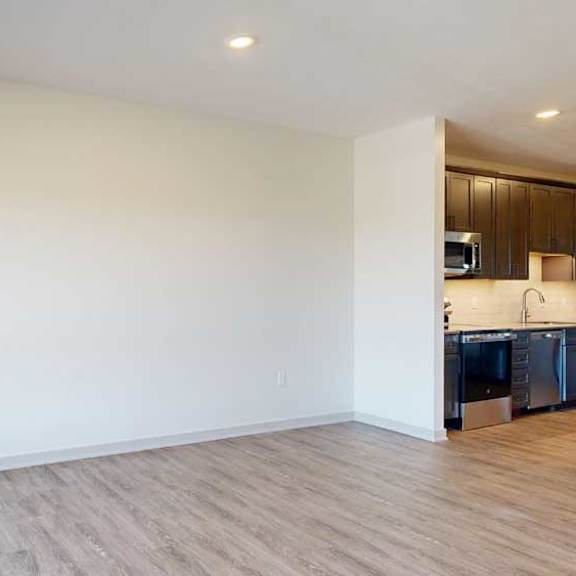 Thumbnail 12 of 21 A living room with hardwood-style flooring and kitchen with dark wooden cabinets.