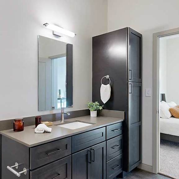 Thumbnail 20 of 23 A bathroom with a quartz vanity, dark wooden cabinets, a tall storage cabinet, and a door open to a bedroom.