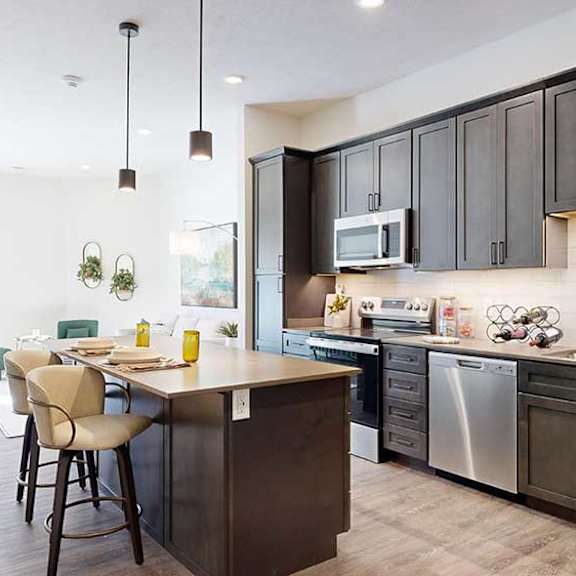 Thumbnail 2 of 23 A kitchen with dark wooden cabinets and a large island, in front of a spacious living room with floor-to-ceiling windows.