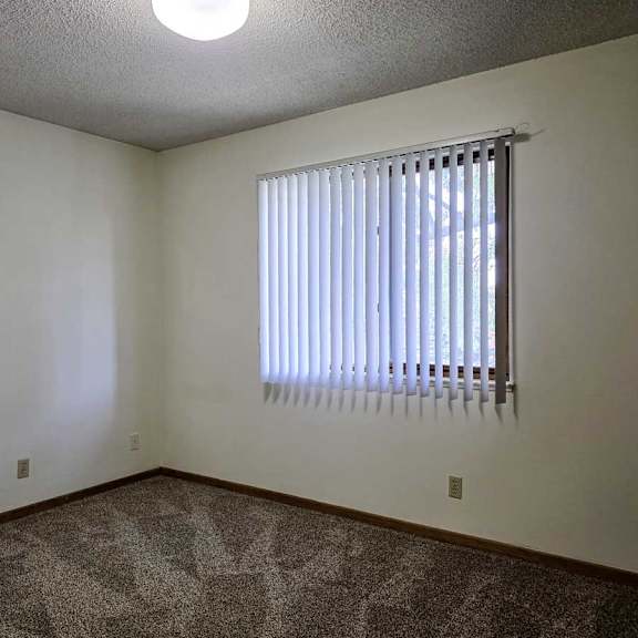 Thumbnail 5 of 11 A carpeted bedroom with a window covered by white vertical blinds.