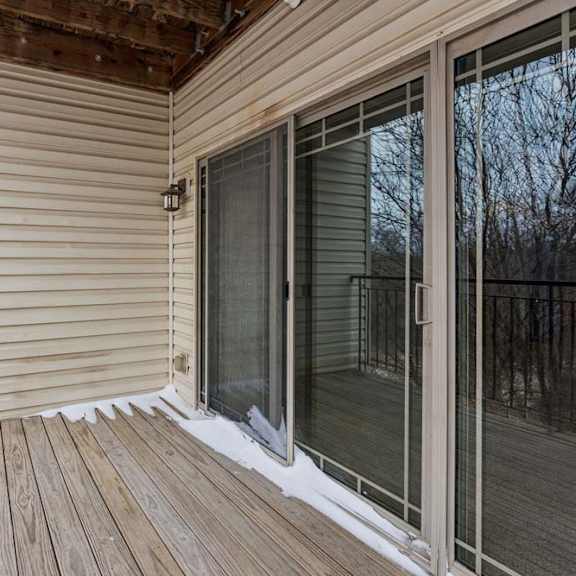 Thumbnail 25 of 26 Private apartment balcony with wood decking, metal railing, and sliding glass doors, overlooking a wooded view with trees in the background.