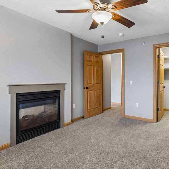 Thumbnail 18 of 26 Apartment living room with neutral carpet, a corner fireplace, ceiling fan, and an open doorway leading to a laundry area with shelving.