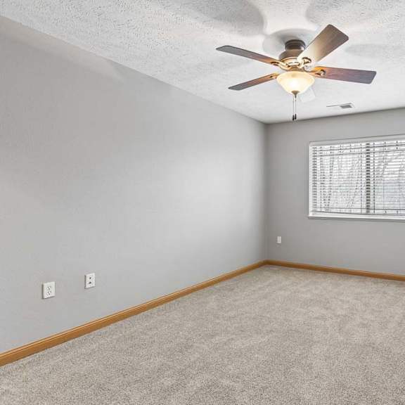 Thumbnail 20 of 26 Bright apartment living room with neutral carpet, a corner fireplace, ceiling fan, and a large window with blinds that provides natural light.