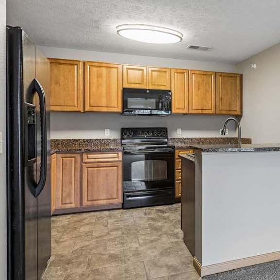Thumbnail 16 of 26 Open-concept apartment kitchen with wood cabinets, black appliances, laminate countertops, and a breakfast bar connecting to the adjacent living area.