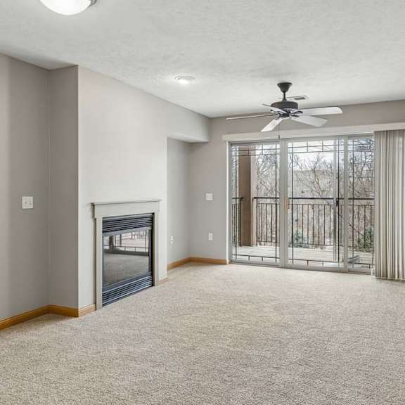 Thumbnail 19 of 26 Spacious apartment living room with neutral carpet, a corner fireplace, and sliding glass doors that open to a private balcony with wooded views.