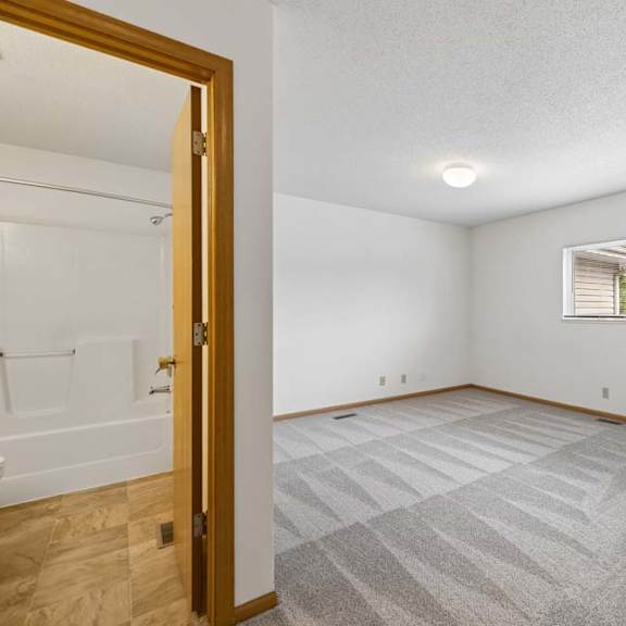 Thumbnail 33 of 37 Bright, empty apartment bedroom with fresh carpeting, a large window, neutral walls, wood trim, and an attached bathroom.