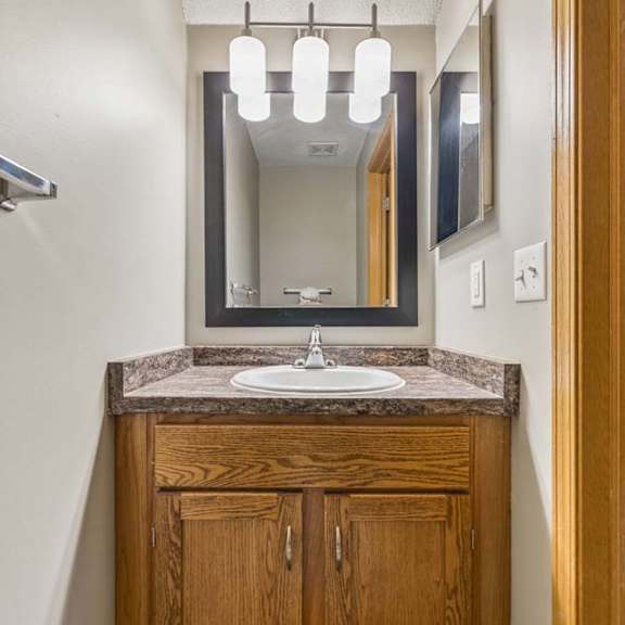 Thumbnail 24 of 26 Compact apartment bathroom vanity with a wood-tone cabinet, laminate countertop, framed mirror, modern light fixture, and towel bar in a neutral interior.