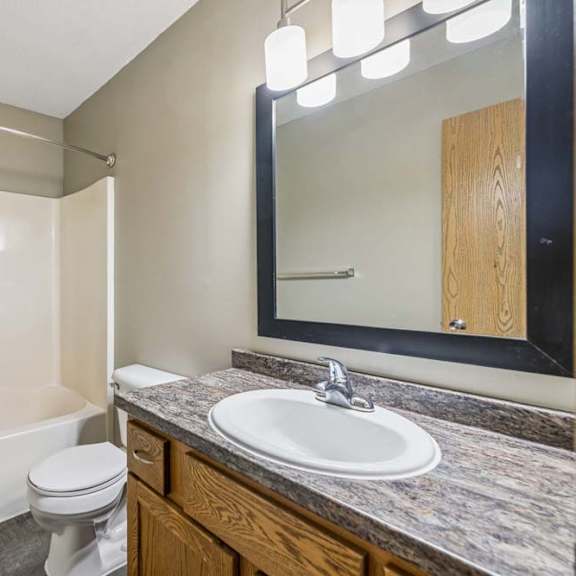 Thumbnail 23 of 26 Apartment bathroom with a single-sink vanity, wood-tone cabinet, framed mirror, modern light fixture, toilet, and a tub/shower combination with built-in shelving.