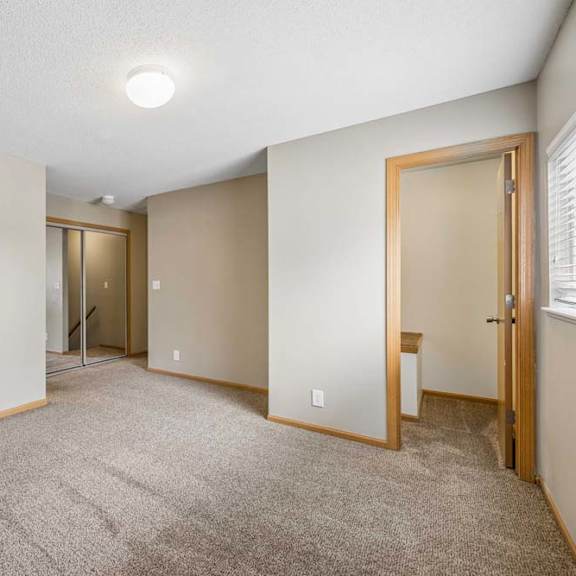 Thumbnail 19 of 26 Bright, empty apartment bedroom with neutral carpet, beige walls, wood-trimmed doors, a window for natural light, and an open doorway leading to the closet area.