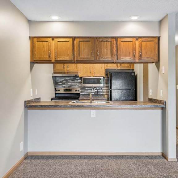 Thumbnail 15 of 26 Open apartment kitchen with a breakfast bar, wood-tone cabinets, black appliances, laminate countertops, and a view into the adjacent living area and hallway.