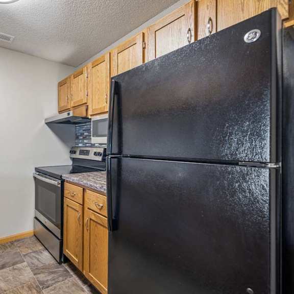 Thumbnail 13 of 26 Galley-style apartment kitchen with wood-tone cabinets, black refrigerator and oven, laminate countertops, and durable flooring in a functional layout.